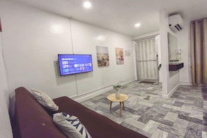 A living room with a flat-screen TV mounted on the wall, a sofa with cushions, a small round table with a vase, and a patterned floor. - VG Pension and Residences by Zuzu (Cebu City)