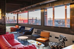 Un espace de détente moderne, doté de sièges et de tables, de grandes fenêtres offrant une vue sur la ville et d’un plafond haut avec des poutres apparentes.