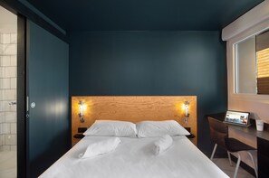 A hotel room with a bed, two bedside lamps, a desk with a computer, and a window with blinds. - Eklo Clermont Ferrand Centre Gare (Clermont-Ferrand)