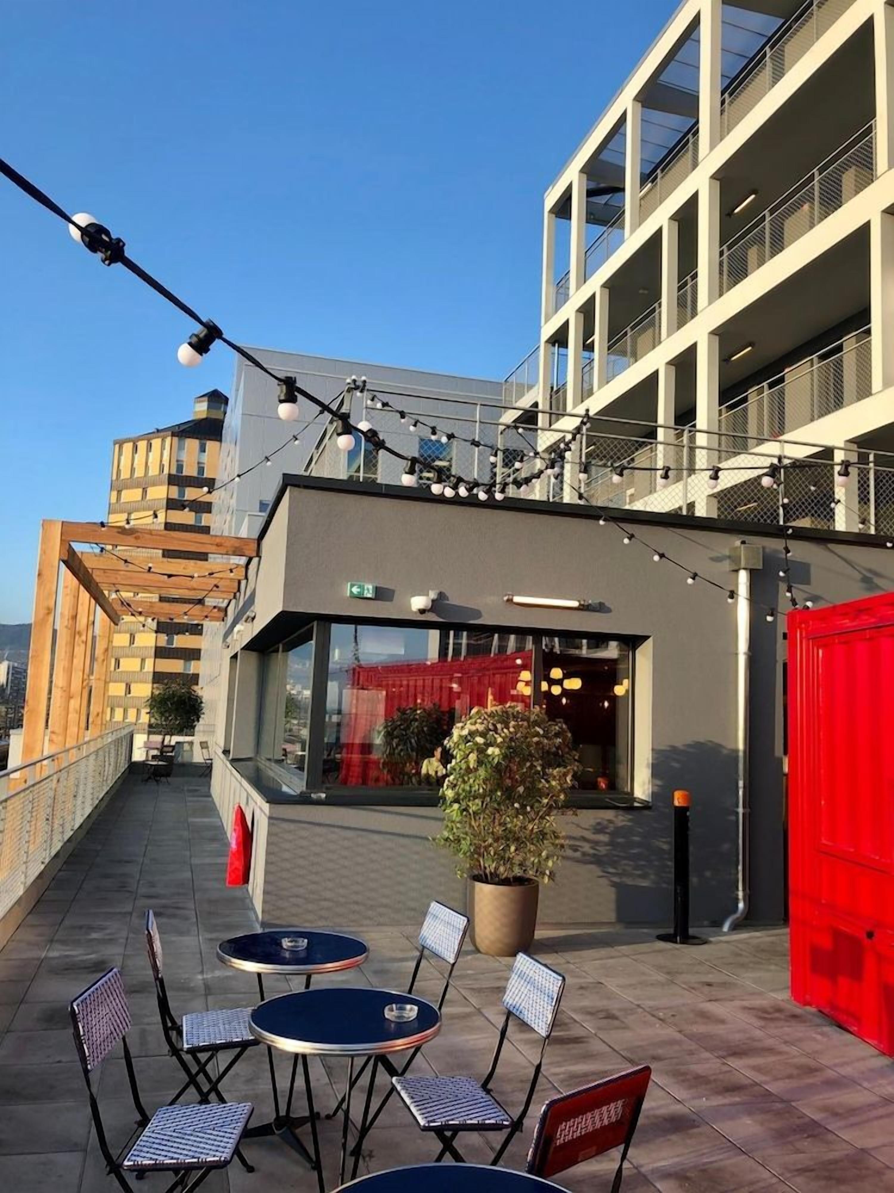 An outdoor seating area with tables and chairs, a potted plant, and a building with balconies and a red door.