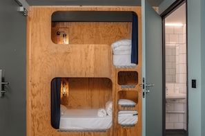 Ein kleines Zimmer mit einem Etagenbett, einem kleinen Fenster und einem durch eine geöffnete Tür sichtbaren Badezimmer.