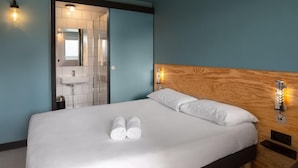 Hotellihuone, jossa on sänky, puinen pääty, yöpöytä, seinään kiinnitetty valaisin ja kylpyhuone, joka näkyy avoimesta ovesta.