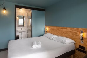 Habitación de hotel con cama, cabecera de madera, mesita de noche, luz adosada a la pared y baño visible a través de una puerta abierta.