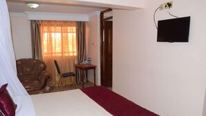 A hotel room with a bed, a chair, a small table, a television, and a window with curtains. - Phoenicia Hotel (Kiambu)
