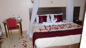 A hotel room with a bed, a chair, a bedside table, and a curtain. - Phoenicia Hotel (Kiambu)