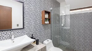 Um banheiro com parede estampada em padrão geométrico, prateleira de madeira com artigos de higiene pessoal, pia branca e vaso sanitário.