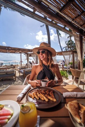 A person seated at an outdoor table with a meal, overlooking a beach and palm trees. - Areia Playa del Carmen (Playa del Carmen)