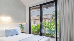 Quarto de hotel com uma cama, mesa de cabeceira, planta em vaso e vista para um edifício e vegetação através de uma porta de vidro.