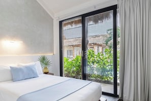 Una camera d'hotel con un letto, un comodino, una pianta in vaso e vista su un edificio e del verde attraverso una porta vetrata.