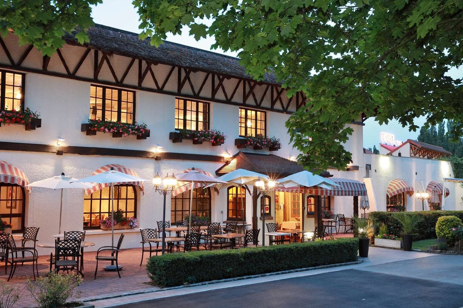 A traditional building with a white facade, dark timber framing, and multiple windows. There are outdoor seating areas with umbrellas and chairs, and a well-maintained hedge in front.