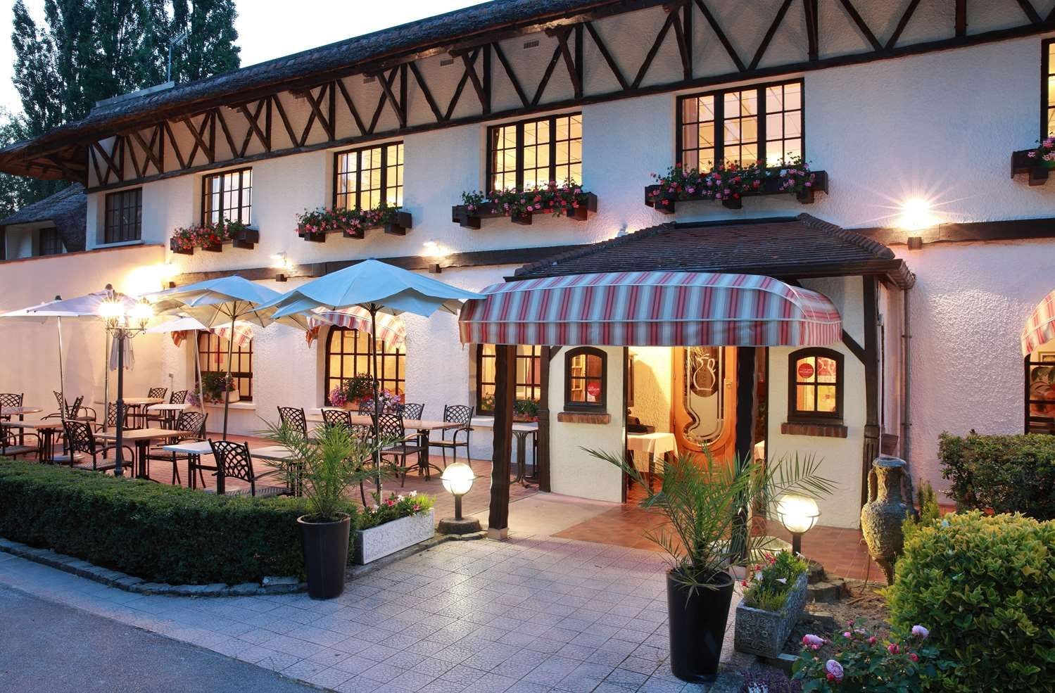 Il s’agit d’un bâtiment traditionnel doté d’un toit de chaume, de nombreuses fenêtres et d’un espace extérieur aménagé avec des tables et des parasols.