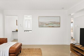 A modern living room with a brown leather sofa, a flat-screen TV on a wooden stand, a large abstract painting on the wall, and a kitchen area visible through an open door. - New Farm Hideaway with Courtyard - 2Bed-1Bath-1Car (New Farm)
