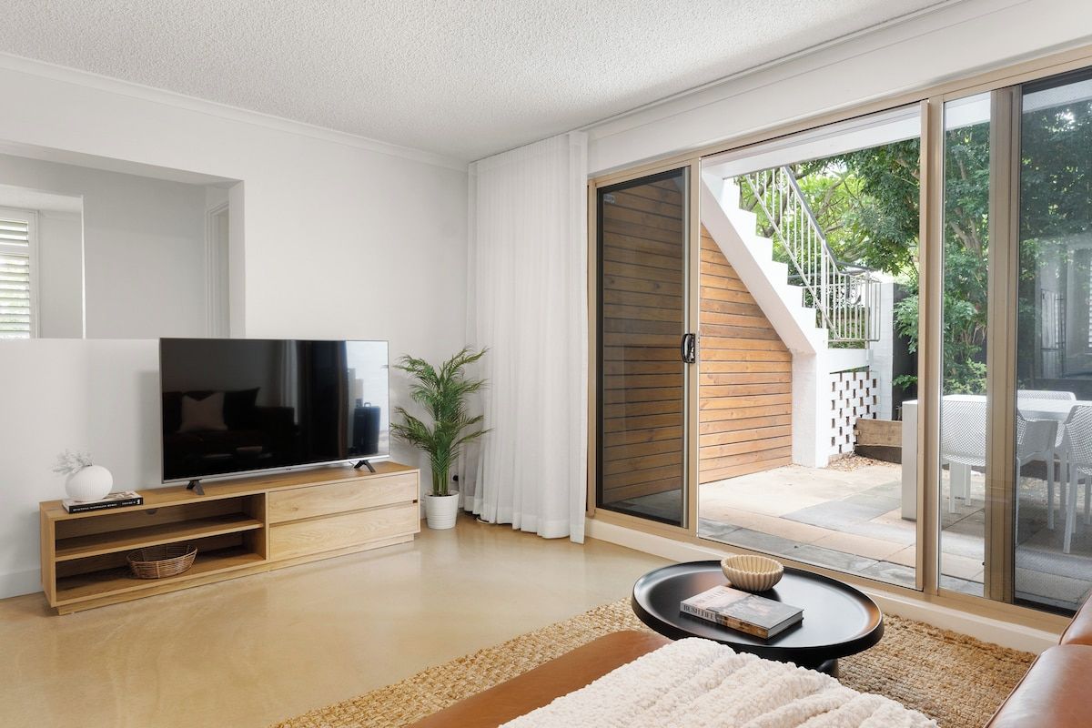 A modern living room with a flat-screen TV, wooden entertainment unit, and a sliding glass door leading to an outdoor patio.