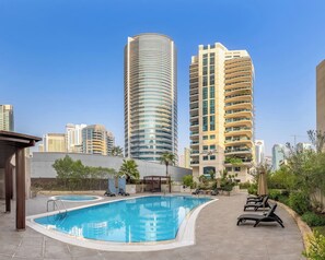 Piscina com espreguiçadeiras, cercada por vegetação e edifícios modernos ao fundo.