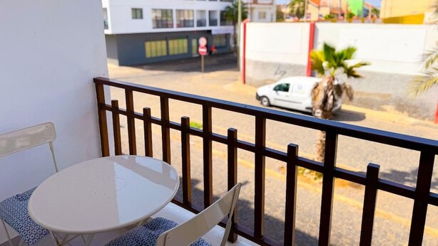 En balkon med et bord og stole, med udsigt over en gade med bygninger og en parkeret bil.