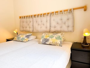 Una cama doble con una colcha blanca y dos almohadones decorativos, un cabecero de madera y lámparas de noche sobre una mesita.