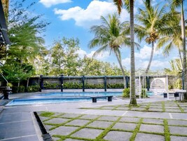 Un área de piscina con un deck de piedra, palmeras y una zona de estar cubierta.