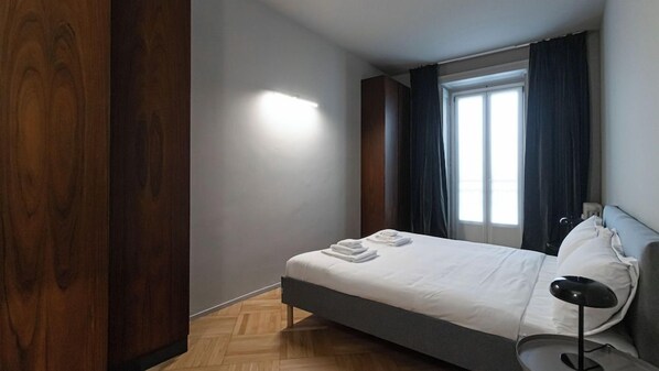 A bedroom with a bed, a nightstand, a lamp, a window with curtains, and a wooden wardrobe. - Italianway Easy - San Vittore 16 (Milan)