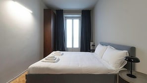 A hotel room with a bed, a bedside table with a lamp, a window with curtains, and a wall-mounted light. - Italianway Easy - San Vittore 16 (Milan)