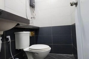 Een badkamer met een toilet, een witte prullenbak en een stopcontact aan de muur.