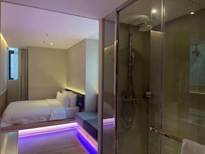 Una habitación de hotel moderna con cabina de ducha de vidrio, cama con ropa de cama blanca y mesita de noche.