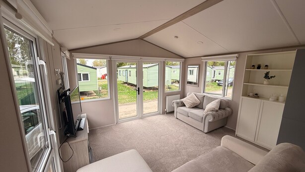 A room with a sofa, a small table, a cabinet with shelves, and a sliding glass door leading to an outdoor area with mobile homes. - Cabin Sleeps 6 With Lake Access Parking (Northampton)