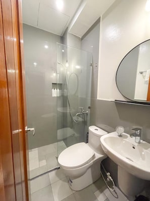 A modern bathroom with a glass-enclosed shower, a white toilet, and a sink with a mirror above. - Koze Suites (Dumaguete)