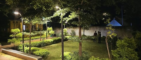 A well-lit outdoor area with a paved walkway, neatly trimmed bushes, and a group of people gathered near a wall.