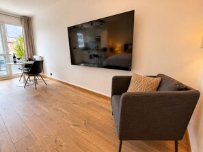 Un salon moderne avec un grand téléviseur à écran plat fixé au mur, un canapé gris agrémenté d’un coussin à motifs et un coin repas comprenant une table et des chaises.