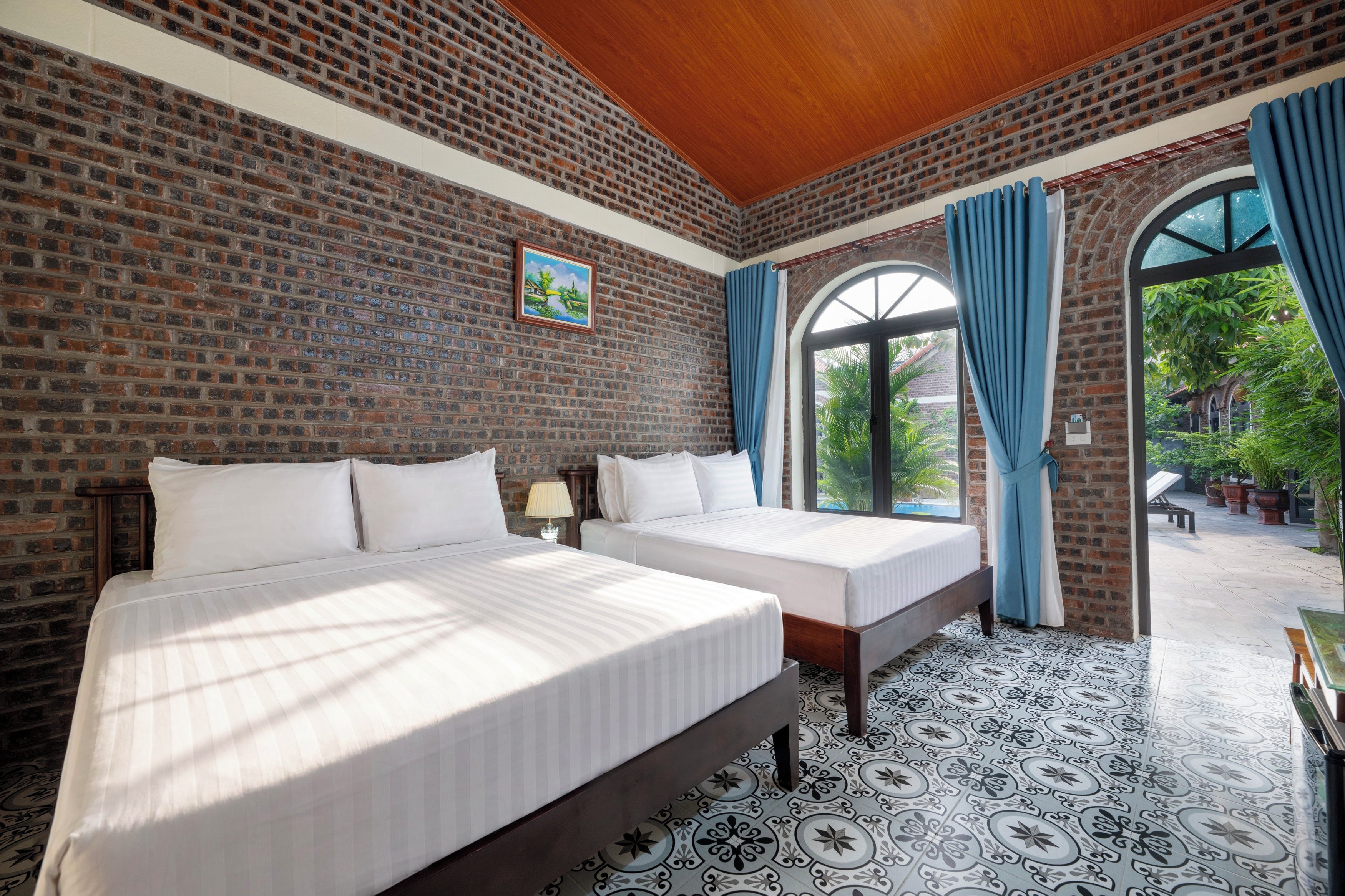 A hotel room with two beds, a brick wall, a large window with curtains, and a view of the outdoors.