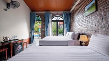 A hotel room with a large bed, a desk with a kettle, a lamp, and a view of a pool and greenery through a glass door.