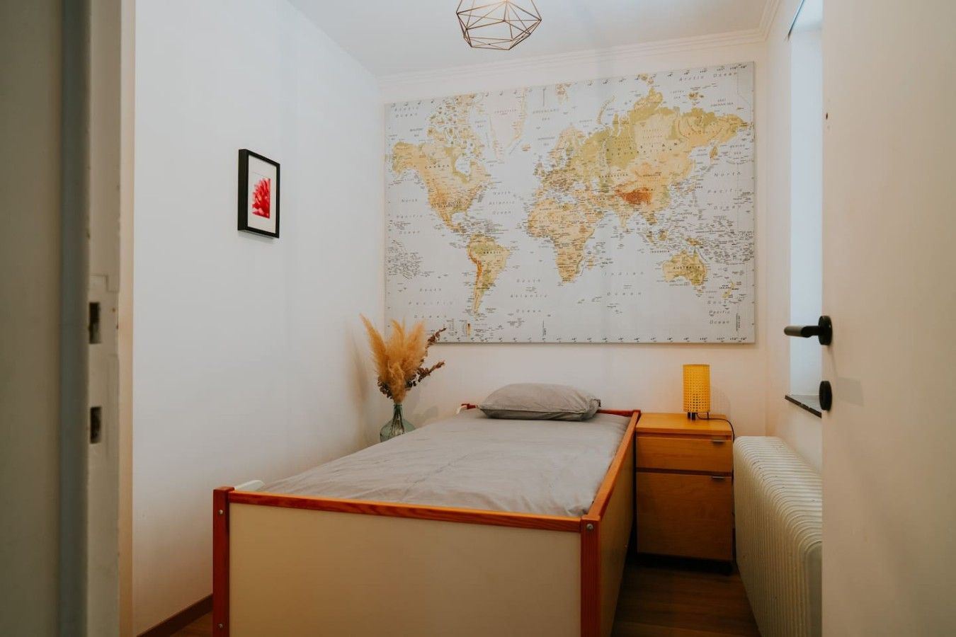 A bedroom with a bed, a nightstand, a lamp, a map on the wall, and a vase with dried plants.