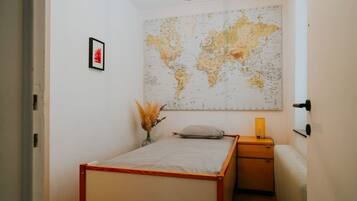 A bedroom with a bed, a nightstand, a lamp, a map on the wall, and a vase with dried plants.