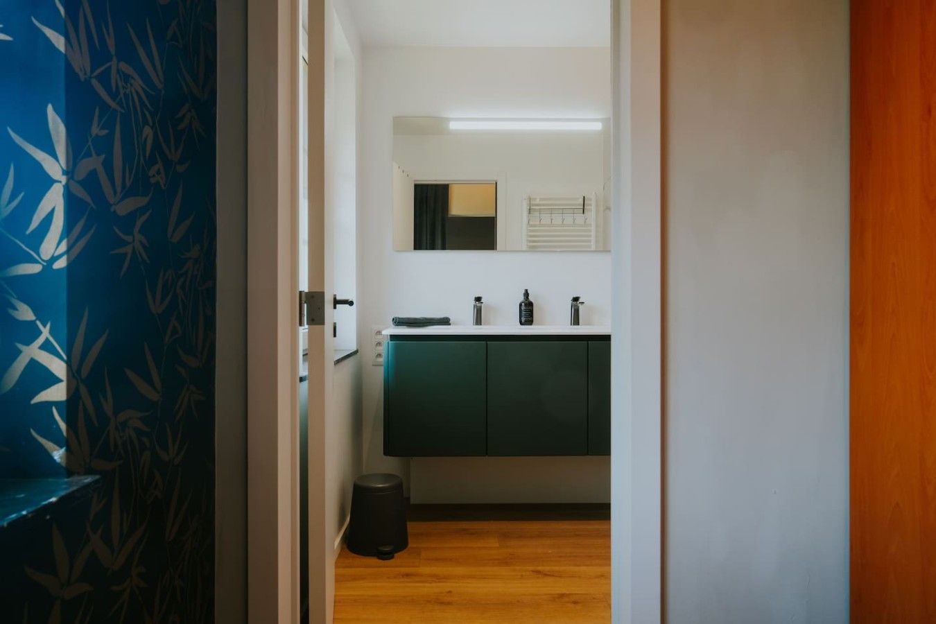 A narrow hallway leading to a bathroom with a white sink, dark cabinets, and a mirror above the mirror.