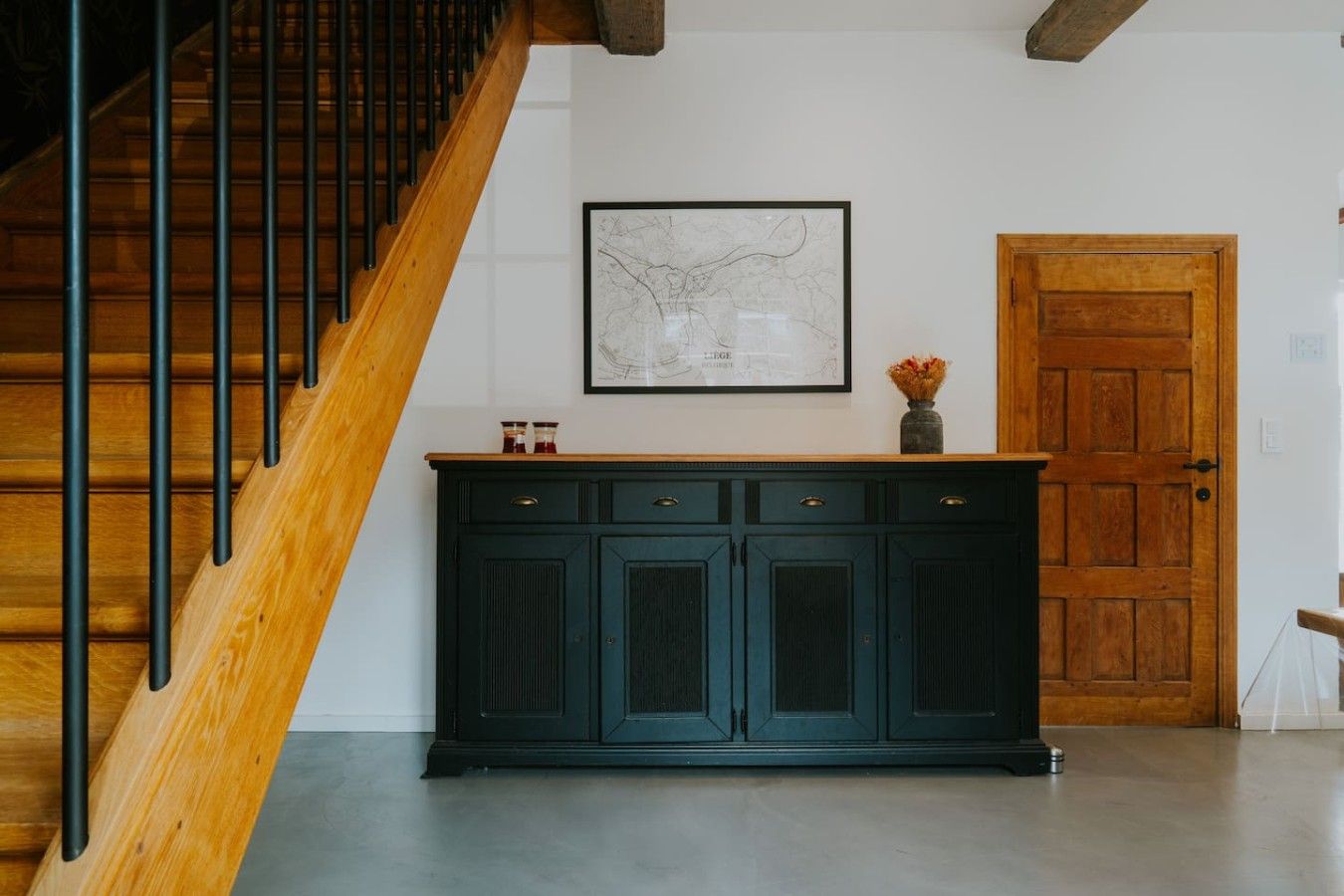 Een trap met houten treden en zwarte metalen leuningen, een donkergroene kast met lades, een inlijsting kaart aan de muur, en een houten deur.