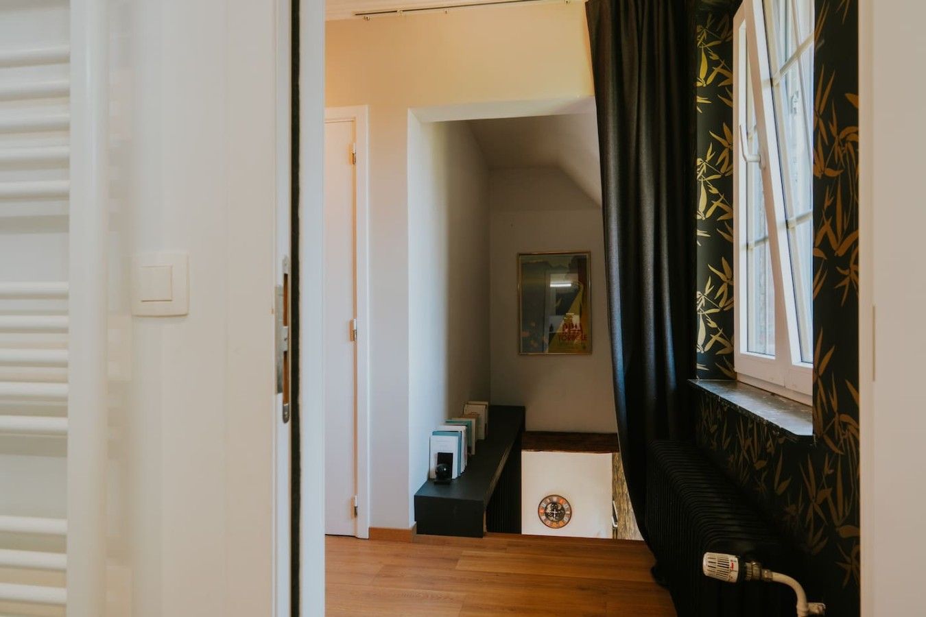 Un couloir avec une porte donnant sur une pièce, une fenêtre ornée d’un motif en bambou et un radiateur fixé au mur.