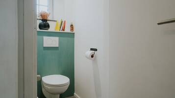 A small bathroom with a toilet, a shelf with books and a vase, and a toilet paper holder on the wall.