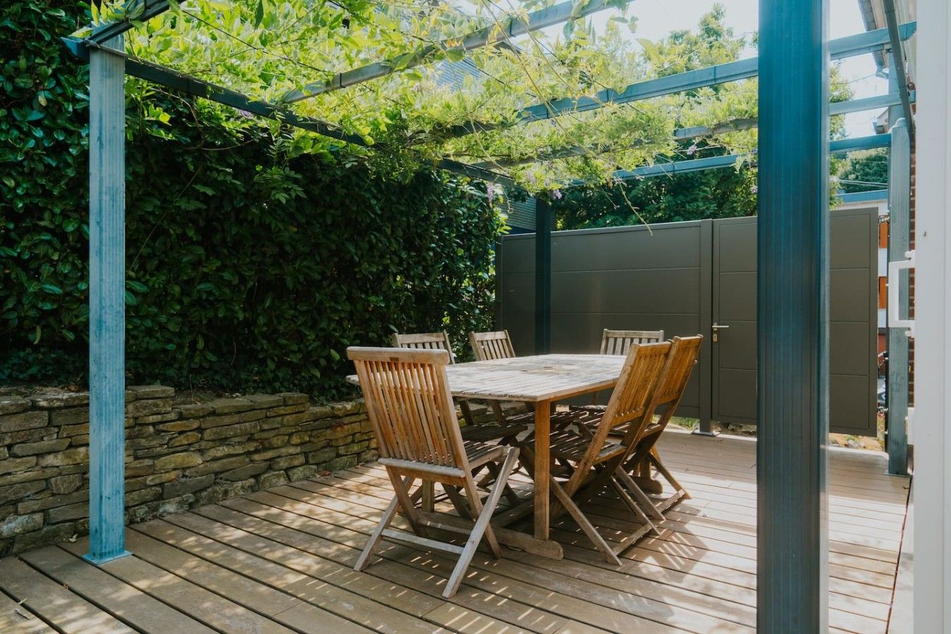 A wooden deck with a table and chairs, surrounded by a stone wall and greenery.