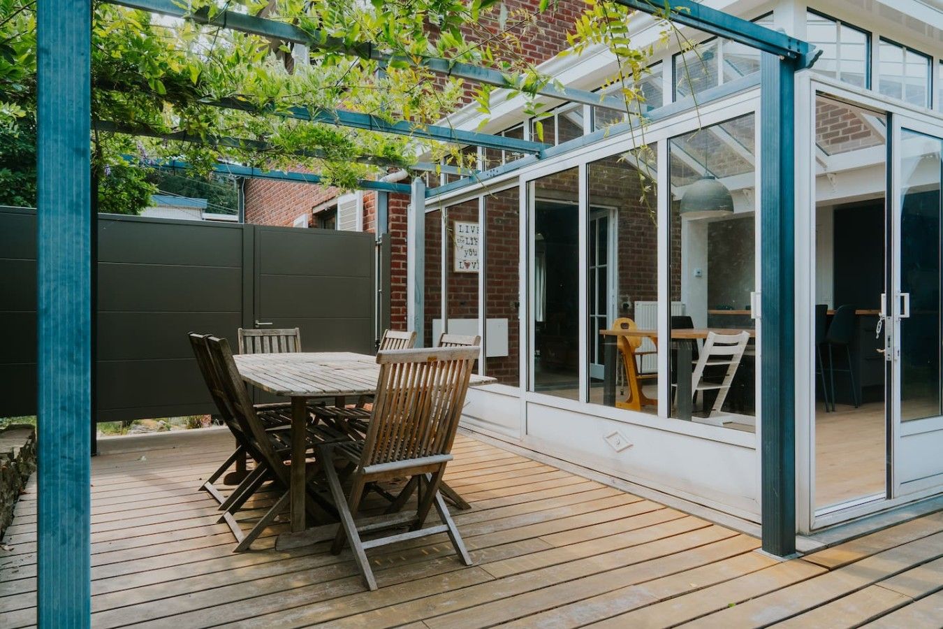 Een overdekt terras met een houten tafel en stoelen, een glazen omheining en een bakstenen muur op de achtergrond.