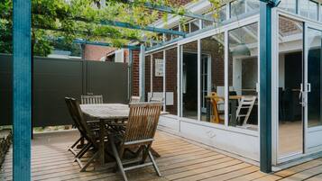 Een overdekt terras met een houten tafel en stoelen, een glazen omheining en een bakstenen muur op de achtergrond.