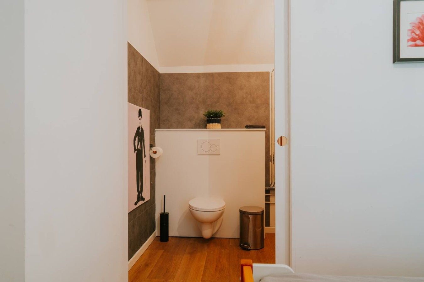 Een kleine badkamer met een toilet, een prullenbak, een toiletrolhouder en een lijstje aan de muur.
