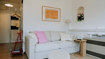 A modern living room with a white sofa, a small round table, a red chair, a metal shelving unit, and a framed artwork on the wall.