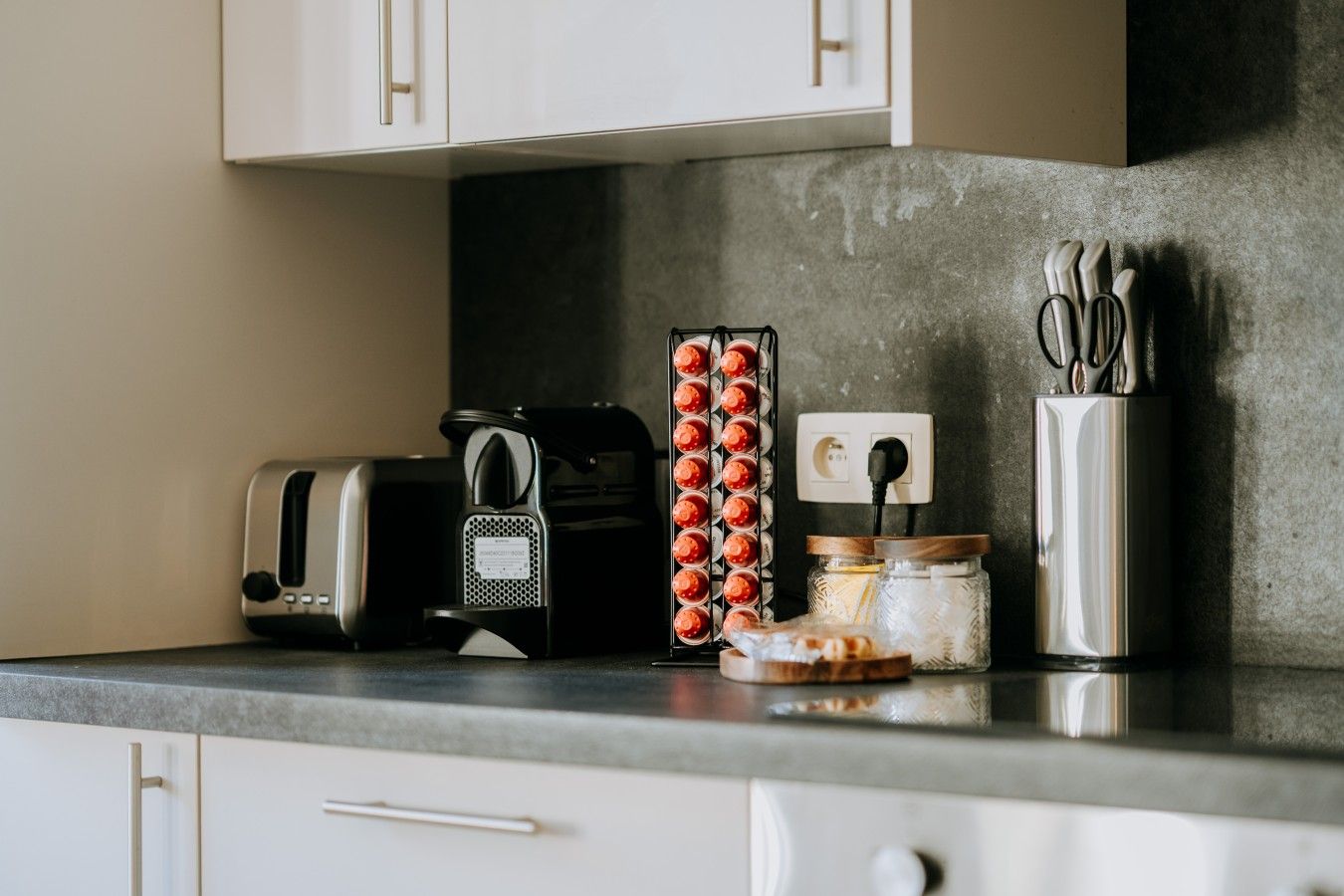 Un plan de travail de cuisine avec un grille-pain, une machine à café, un pot de confiture, un récipient contenant des ustensiles et une prise de courant.