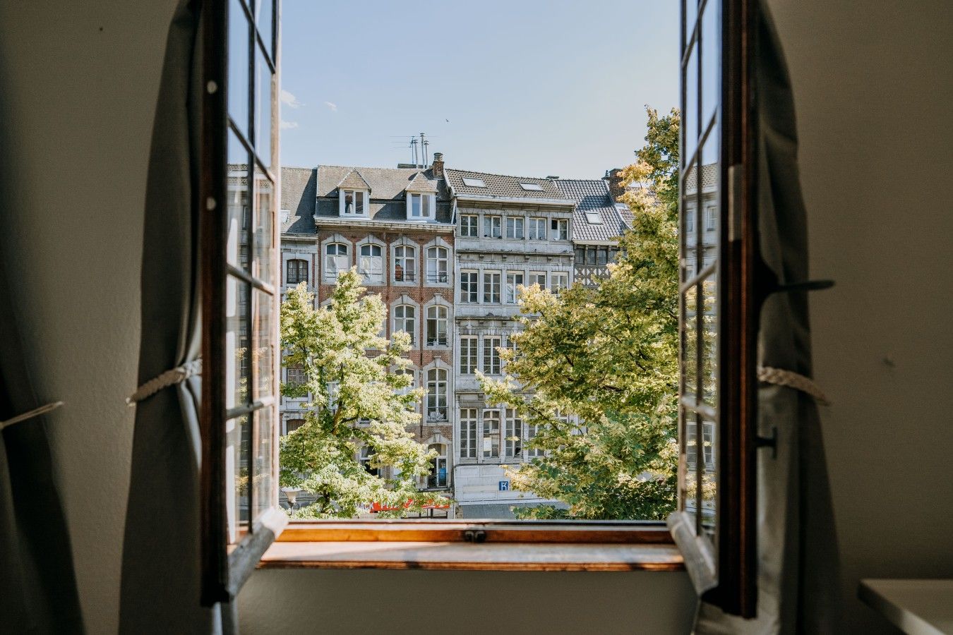 Une vue à travers une fenêtre dotée d’un rebord en bois, donnant sur un immeuble à plusieurs étages de style architectural traditionnel, caractérisé par un mélange de façades en brique et en pierre, et entouré d’arbres.