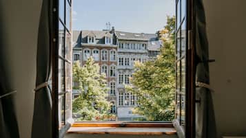 Une vue à travers une fenêtre dotée d’un rebord en bois, donnant sur un immeuble à plusieurs étages de style architectural traditionnel, caractérisé par un mélange de façades en brique et en pierre, et entouré d’arbres.