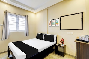 Quarto de hotel com cama, mesa de cabeceira, escrivaninha e um vaso de flores, e janela com cortinas.