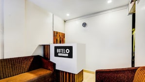 Zona de recepción de hotel con mostrador identificado como 'HOTELO Company Serviced', un reloj en la pared y dos sofás.