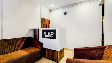 Zona de recepción de hotel con mostrador identificado como 'HOTELO Company Serviced', un reloj en la pared y dos sofás.