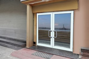 Une entrée d’immeuble avec un volet métallique fermé, un escalier menant à une porte, et une vue sur un bâtiment éloigné, visible à travers la porte.