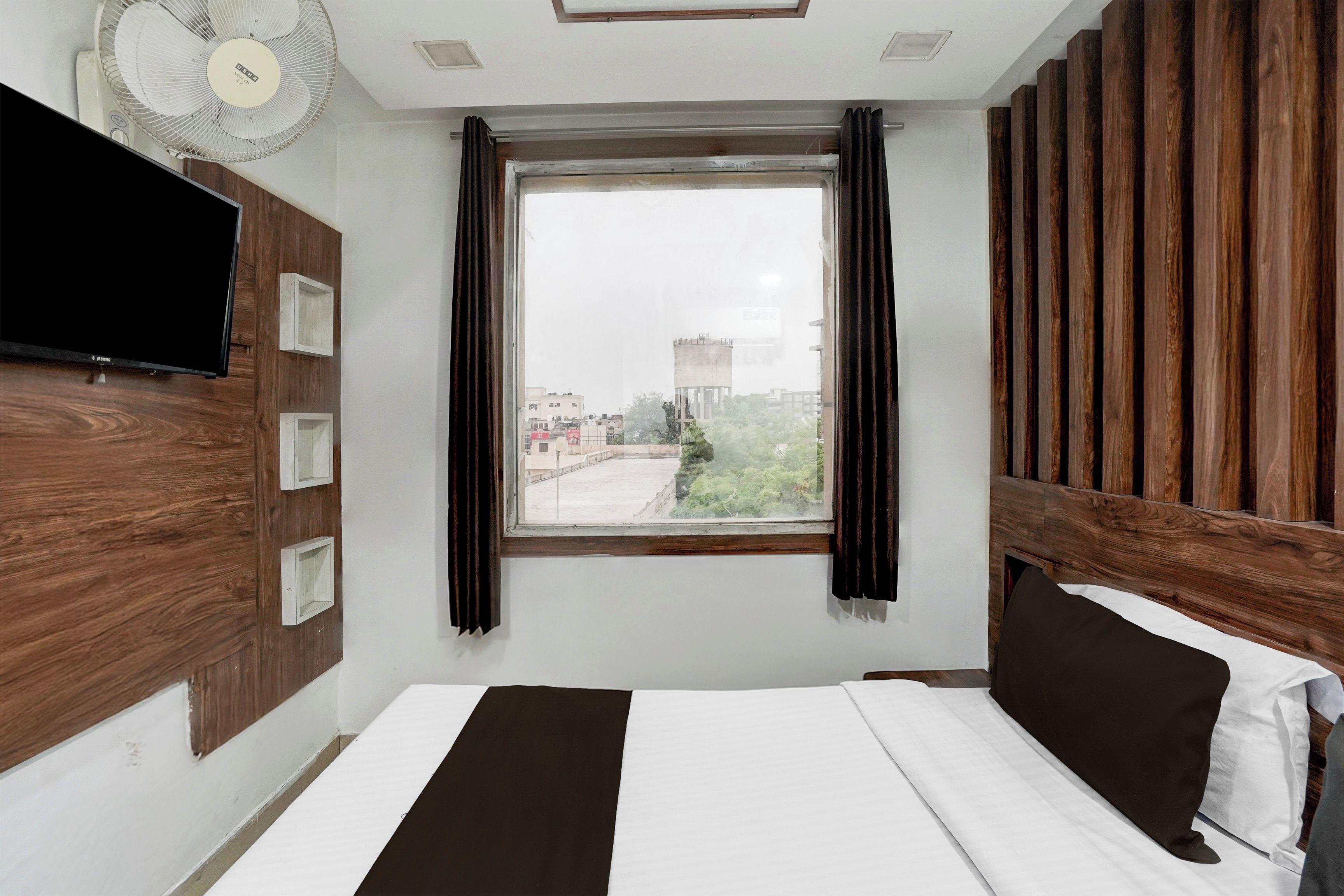 A hotel room with a bed, a TV, a window with curtains, and a view of the cityscape.
